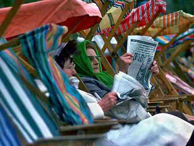 two women in deck chairs at seaside reading newspapers